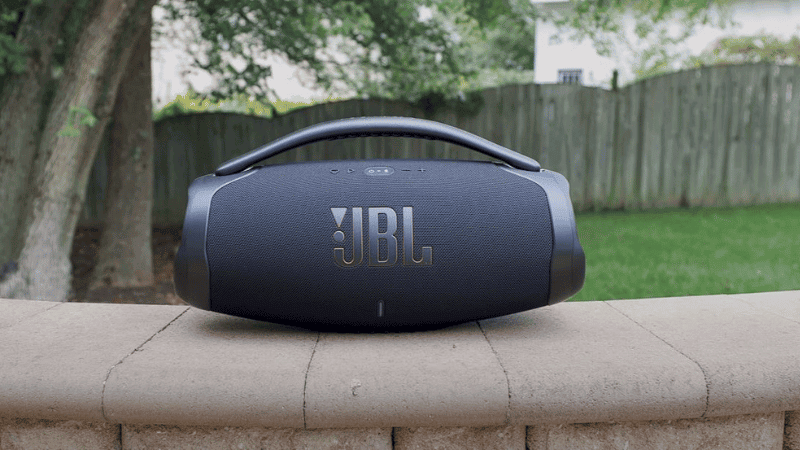 Vue de face, au premier plan, d'une enceinte portable JBL Boombox 3 Wi-Fi posée sur un mur en pierre, avec un arbre, un jardin et une barrière en bois en arrière-plan.