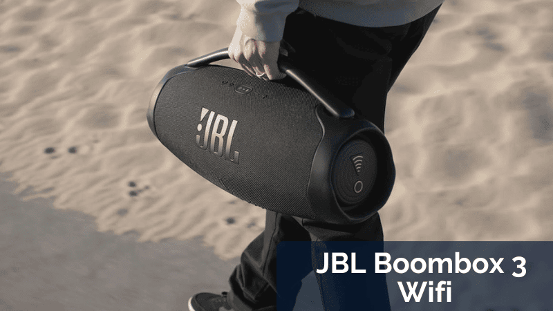 Vue du dessus d'un homme portant une JBL Boombox 3 Wi-Fi dans sa main gauche. L'homme est sur son skateboard et se déplace sur la plage de sable, en direction de la mer